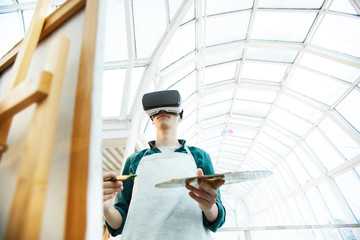 Low angle portrait of young man wearing VR headset painting picture on easel under futuristic glass...