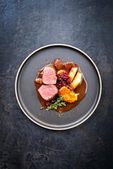 Traditional saddle of venison with fried mashed potatoes and fruits in game red wine sauce as top view on a modern design plate with copy space