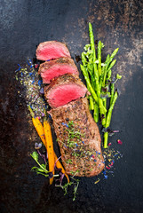 Traditionell gegrillter Hirschrücken mit grünen Spargel, Karotten und Kräuter als Draufsicht auf einem alten rustikalen Blech © HLPhoto