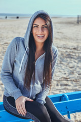 Portrait of a cheerful beautiful girl with tanned skin wearing a gray hoodie sitting on a bench at the beach
