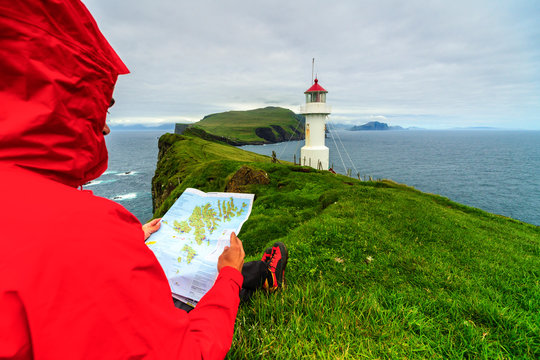 Hiker on cliffs looks at the map next to lighthouse, Mykines island, Faroe Islands, Denmark