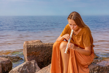 Young girl with long blond hair in long dress playing flute sitting on stone on sea shore © Andzo Janovich