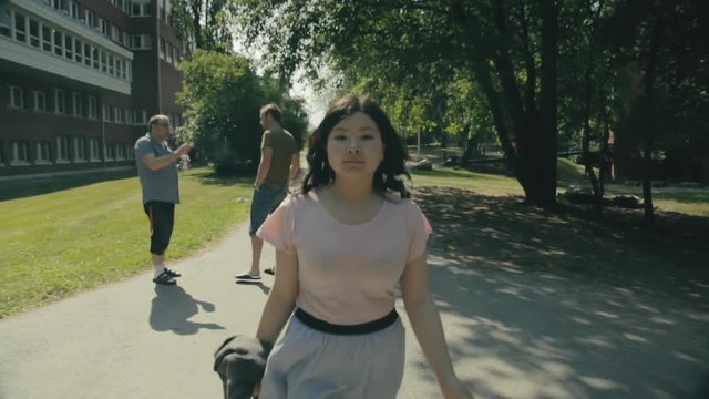 A young woman in a skirt walks down a path on campus past two men talking outside a dormitory building. Pretty Chinese lady walking outside on a sunny afternoon.