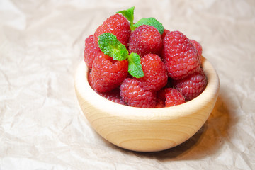 A wooden bowl with juicy delicious red raspberries decorated with fresh mint on the plain bright background