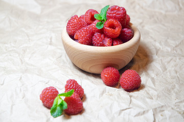 A wooden bowl with juicy delicious red raspberries decorated with fresh mint on the plain bright background
