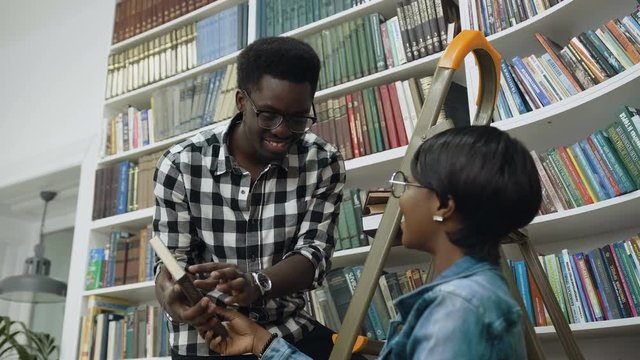 African guy standing on ladder and giving books to african girl in university library.