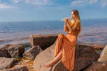 Young girl with long blond hair in long dress playing flute sitting on stone on sea shore