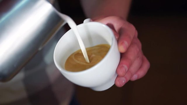 Barista pouring latte foam over coffee making a perfect cappuccino and creating a white heart. White cup with cappuccino