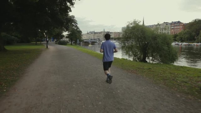 Wide follow shot of an Asian man jogging along a waterway with calm water and sailboats lined up on the opposite bank. Classic architecture of Stockholm seen in the buildings along the river.