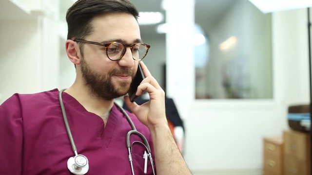  Young doctor in glasses talking on the phone in a purple medical uniform in the office.