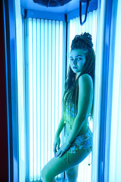 Solarium. Young pretty girl with african braids in a dress for oriental dances sunbathing in a vertical sunbed. Blue neon glow.