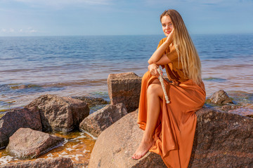 Young girl with long blond hair in long dress holding flute sitting on stone on sea shore © Andzo Janovich