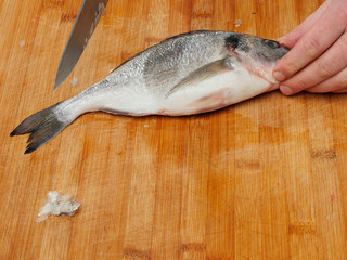 Cleaning raw sea bream on a wooden table with a knife. Hand holding fish by the head.