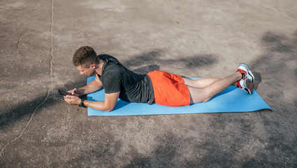 A male athlete, in the summer in the city, lies on the athletic mat and looks at smartphone....