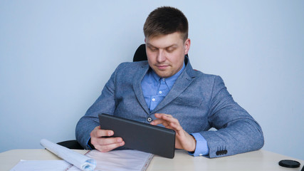Man (guy) An office worker happily looks at the tablet laughing from what he sees on the screen. Concept of: Happy workday, Architecture, Hard hat, Paper, Workday, Lifestyle.