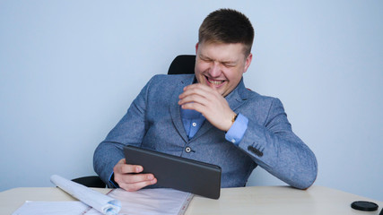Man (guy) An office worker happily looks at the tablet laughing from what he sees on the screen. Concept of: Happy workday, Architecture, Hard hat, Paper, Workday, Lifestyle.