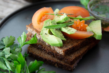 Healthy avocado sandwich on dark rye toast bread made with fresh sliced avocado, seeds and tomatoes on rustic wooden background.