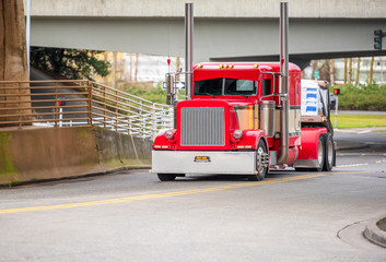 Big rig bright red classic American idol semi truck for long haul routs with high chrome exhaust pipes transporting semi trailer driving on the city street intersection turning under railroad bridge