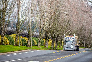 Fototapeta na wymiar Classic big rig American semi truck with flat bed semi trailer transporting heavy equipment on the road with trees alley