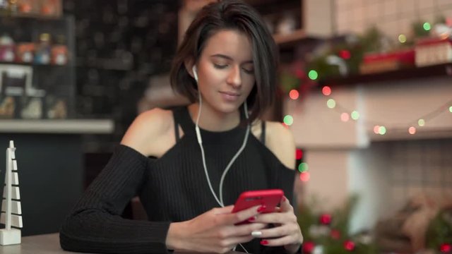 Sexy brunett girl in black shoulderless sweater leaning to the table in cozy cafe, listening to music in earphones, dancing in head, looking at/using phone. Blurred cafe background