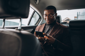 Young smiling african man using smartphone while sitting on backseat in car. Concept of happy...