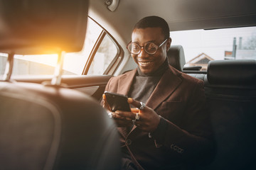 Young smiling african man using smartphone while sitting on backseat in car. Concept of happy business people traveling.