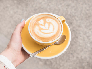 Hot Cappuccino In Yellow Cup On the city street background. The Latte Art. Happy morning time. Female hand holding mug with hot beverage.