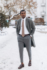 Portrait of handsome African - American businessman using smartphone in snowy winter city street, typing text message and looking at screen.