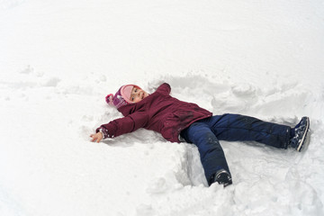 Fototapeta na wymiar Little cute girl in winter outdoors. She makes snow angel. Girl rejoices and fools