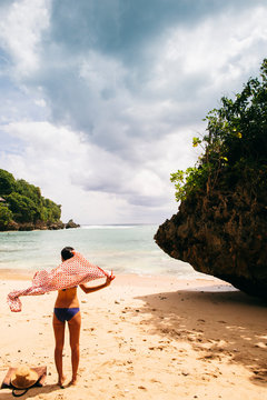 Woman sitting on a beach in a bikini, wrapping on a flowing sarong faces out to the sea towards tropical waters.