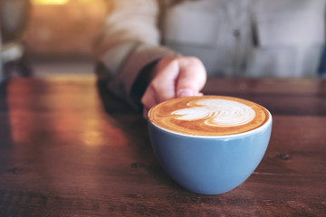 Closeup image of a hand holding a blue cup of hot latte coffee with latte art on wooden table in...