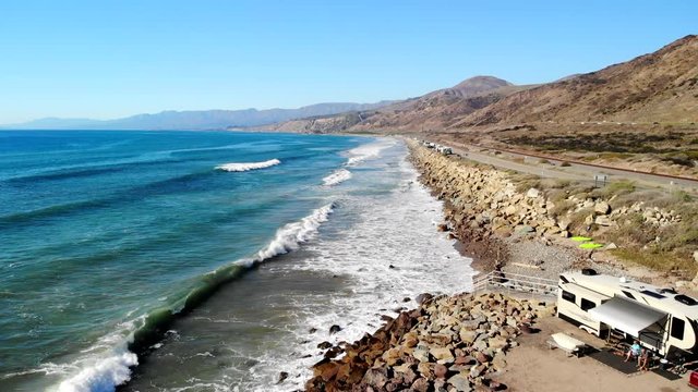 Aerial drone shot rising over an rv campground on the beach with blue ocean waves crashing against the golden California coast line.