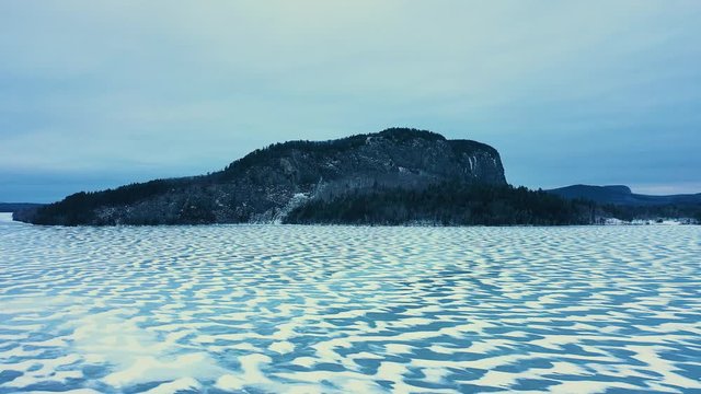 Aerial SLIDE over a frozen lake with snowy patterns past an isolated mountain with a steep cliff face