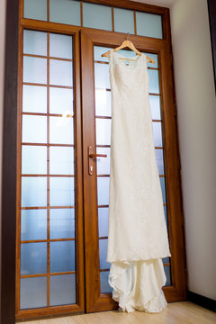 Wedding Dress Hanging On The Background Of A Wooden Door. Wedding. Fees Bride. Home Interior. Door With Stained Glass. White Wedding Shoes.