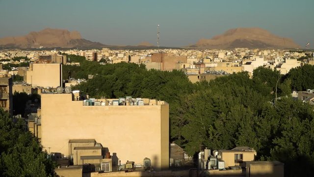 A wide still shot of a sprawling Middle Eastern city, with trees and a mountainous background. Beige apartment rooftops covered in air conditioning units.
