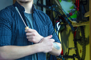 Bearded Classy Young Man with a Tattoo in a T-Shirt and Plaid Shirt Prepares To Maintenance Bicycle. Arms Crossed Holding Tools. Bicycles and Skate in the Storeroom. Technical Repair Service Concept.