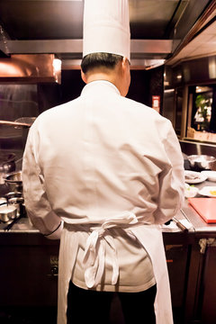 The view from behind of a Chinese chef in tall toque (chefs hat) at his work station.