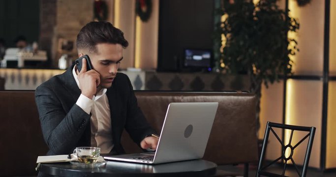 Stressed young businessman with laptop working at cafe. He talking with boss talk phone and writes the notes to the notebook. Hard business, people, deadline and technology concept