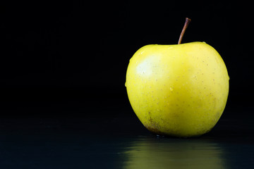 Natural, organic, freshly washed golden delicious apple with the stem on a black stone surface with reflection and a space for text