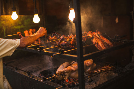 outdoor street chicken barbecue grill arm and hand brush oiling the grilling meat food broiler spit roasted over smoking living coals in a simple electric illumination, the Philippine lechon Manok