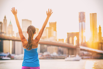 Happy cheering fit woman achieving fitness goal in New York City with arms up at sunset and...