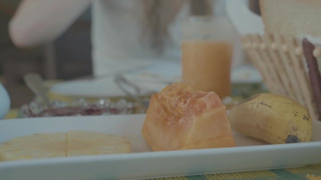 Panning around a plate of tasty food includinng a banana, pineapple slice and a jelly like traditional egg cake at a breakfast table with blurred background of a person and juice