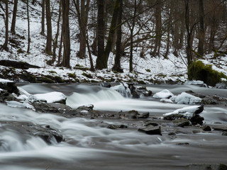 Fototapeta na wymiar smooth motion of wild water in a river in winter with snow and ice on rocks and stones in the beautiful nature of a forest