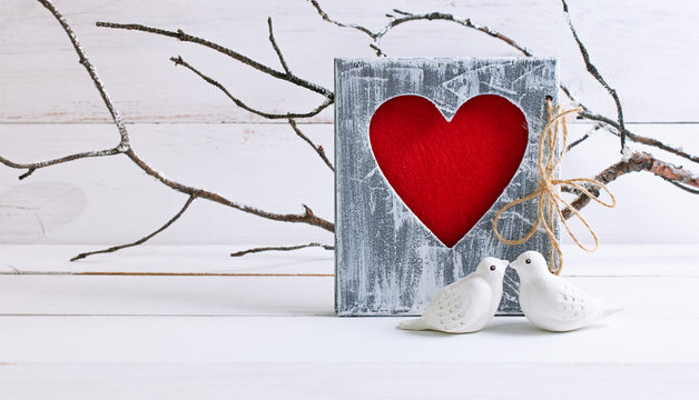 Home decor for Valentines Day with figurines of kissing pigeons on background of branches covered with hoarfrost and shabby chic photo frame with felt heart