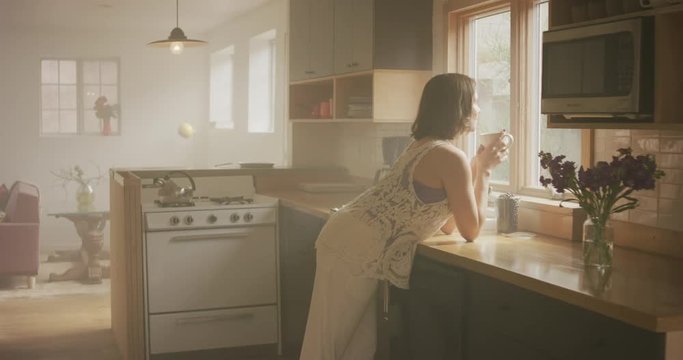 A woman is resting on her kitchen counter looking out the window.  She stands up still gazing into the beautiful sunny day which fis streaming into the room.
