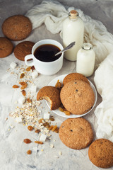 Cup of black coffee in a white cup, Bottles with milk, oatmeal cookies, oatmeal, raisins on a light background. Scene from breakfast
