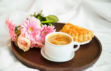 Flowers bouquet with coffee cup and pastry bun on wooden tray in the bed