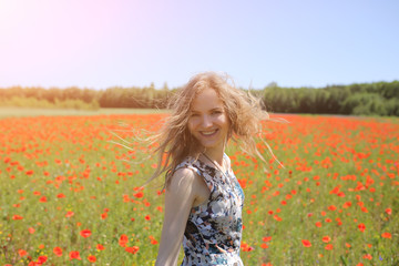 Naklejka na ściany i meble Beautiful girl is standing on summer field full of red poppy flowers in the grass. Sunny day with green lawn background. Happy woman in rustic dress is smiling. The wind in her hair.