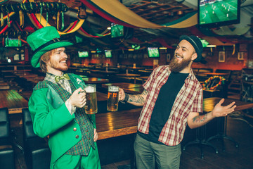 Two friends stand in pub smile. They hold mugs of beer. Guy on left wear saitn patrick's suit. They happy to see each other.
