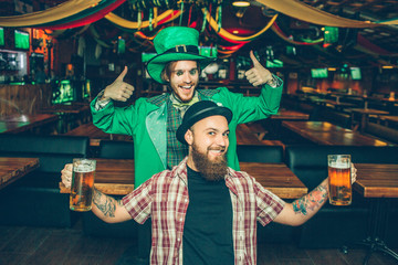 Happy friends stand in pub and pose on camera. They look straight. Guy in green suit show his big thumbs up. Young man in front has two beer mugs in hands.